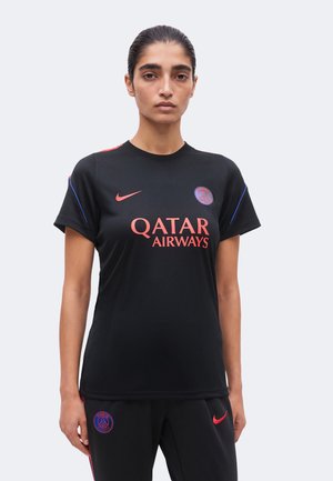 Kobieta o ciemnych włosach w czarnej koszulce piłkarskiej Paris Saint-Germain i dopasowanych spodniach z czerwonymi i niebieskimi akcentami na jednolitym tle.