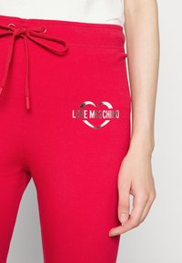 Czerwone dresy mają elastyczny pas ze sznurkiem i metaliczny wzór serca z napisem "LOVE MOSCHINO" w wyraźnej czcionce.