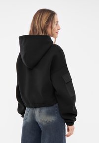 Felpa nera corta con un grande cappuccio, polsini a costine e una tasca laterale, realizzata in un materiale morbido; indossata con jeans blu a vita media.