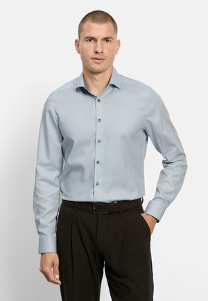 Mann trägt ein hellblaues, langärmeliges Hemd, das in dunkelbraune Hose mit schwarzem Gürtel gesteckt ist, steht mit einer Hand in der Tasche.