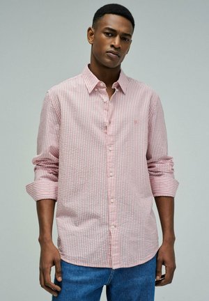 Joven con cabello corto vistiendo una camisa de botones a rayas verticales en rosa y blanco con las mangas remangadas y jeans azules, sobre un fondo gris.