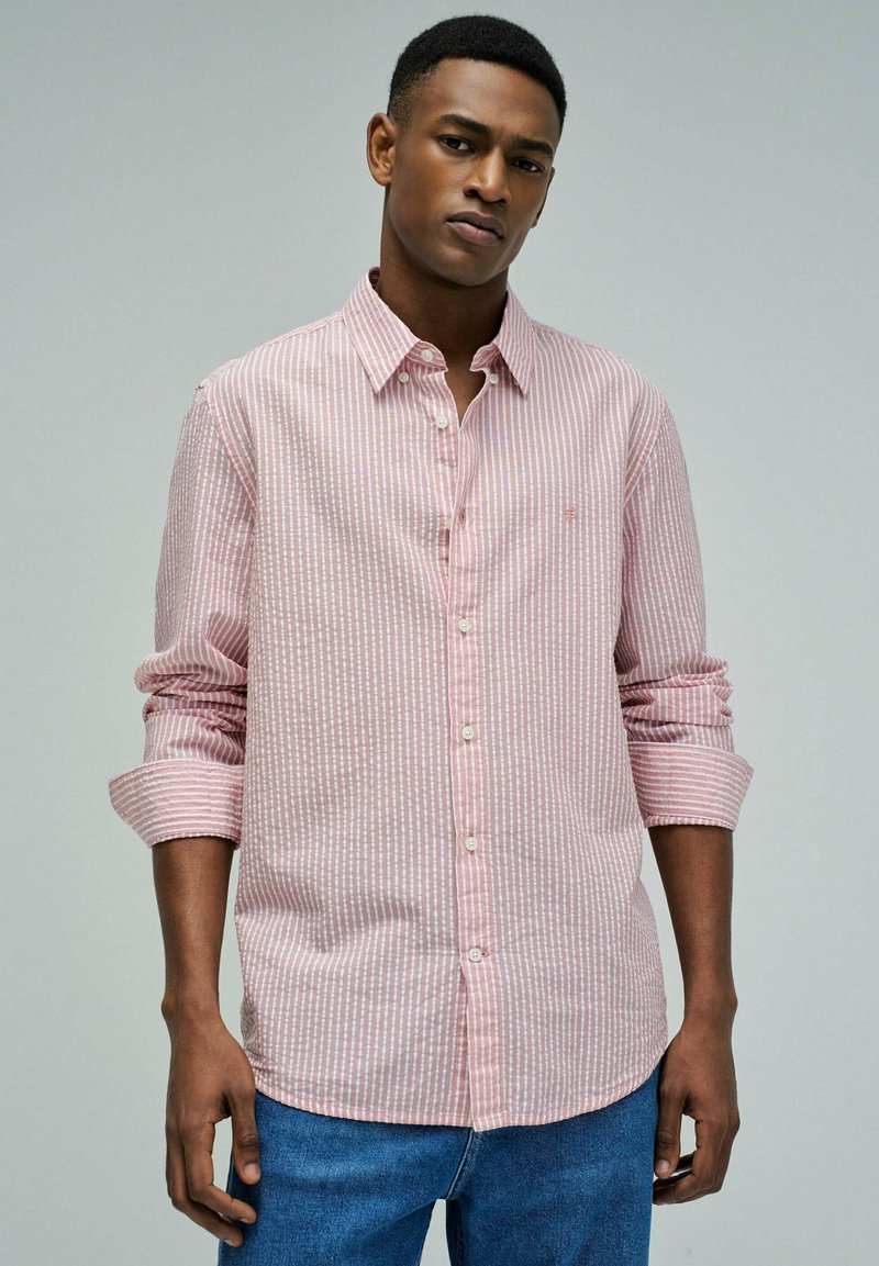 Junger Mann mit kurzen Haaren, der ein rosa-weiß gestreiftes Hemd mit Knöpfen und hochgekrempelten Ärmeln sowie eine blaue Jeans trägt, vor grauem Hintergrund.