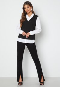 Top senza maniche nera sopra una camicia bianca con colletto, abbinata a pantaloni neri con fessure laterali. Tessuto liscio con un design pulito.
