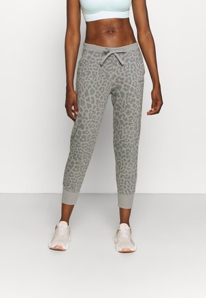 Kobieta ubrana w jasnoszare spodnie dresowe w panterkę, biały sportowy biustonosz i białe trampki, stojąca na tle gładkiego jasnego tła.