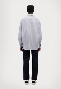 Camisa de rayas azules y blancas de manga larga con cuello abotonado, combinada con jeans de mezclilla oscura y zapatillas claras. Detalles de costura visibles.