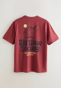 Punainen puuvillateeppari, jossa on kuva surffaajasta ja teksti "Big Wave Society" paksulla mustalla fontilla ja keltaisilla yksityiskohdilla tasaisella taustalla.
