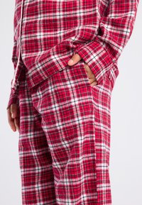 Persoon in rood-wit geruite pyjama met een hand in de broekzak tegen een eenvoudige achtergrond.