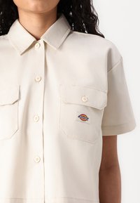 Bežina košulja kratkih rukava na dugme s oštrim ovratnikom, dva džepa na prsima i Dickies logom. Glatka tkanina s suptilnim sjajem.