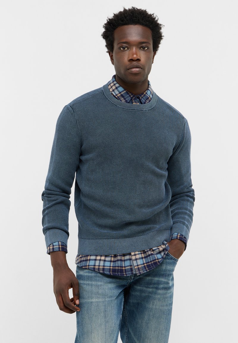Niebieski sweter dzianinowy o teksturowanym wzorze, z okrągłym dekoltem i ściągaczami, noszony na niebieskiej koszuli w kratę z guzikami. W zestawie z jasnoniebieskimi dżinsami.