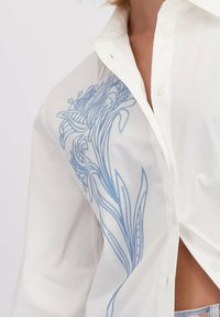 Witte knoopshirt met lichtblauwe abstracte bloemenborduurwerk op de borst, gedeeltelijk open waardoor huid en de tailleband van spijkerbroek zichtbaar zijn.