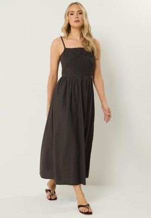 Donna che indossa un abito lungo nero senza maniche con corpetto ricamato a fiori, cammina a piedi nudi su uno sfondo chiaro e semplice.