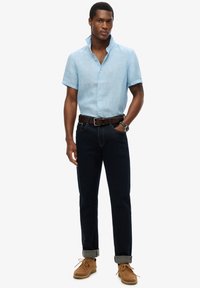 Hombre con camisa de manga corta azul claro, vaqueros oscuros con puños, cinturón marrón, reloj y mocasines color canela, de pie con una mano en el bolsillo.