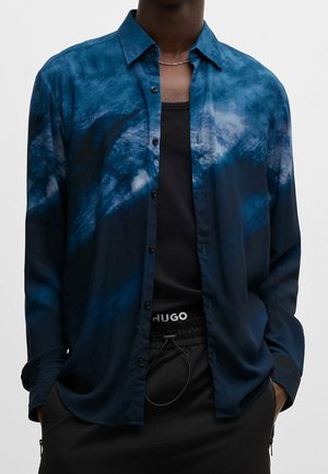 Man met een blauw-zwart tie-dye overhemd open gedragen over een zwart tanktopje en zwarte broek, met een zichtbare witte "HUGO" tailleband.