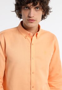 Chemise orange clair à boutons en tissu au toucher doux ; elle présente un petit logo jaune sur la poitrine et des détails en boutons.
