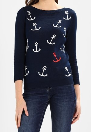Pull bleu marine avec un motif d’ancre blanc et rouge, porté avec un jean bleu foncé, montrant le torse et les bras.