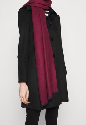 Personne portant un manteau noir boutonné et une longue écharpe bordeaux sur un haut rayé noir et blanc, sur un fond uni clair.