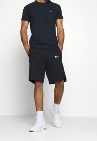 Marineblå idrottströja med raglanärmar, parat med svarta shorts med sidoventiler. Vita varumärkesdetaljer på båda plaggen. Vita sneakers.