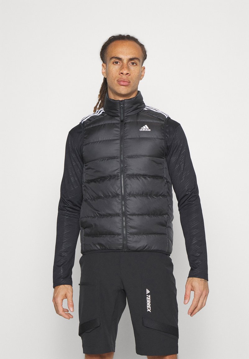 Crna puf jakna bez rukava s visokim ovratnikom, prednjim patentnim zatvaračem i bijelim Adidas logom na prsima. Ima horizontalnu kviltanu površinu i kontrastni materijal na rukavima.