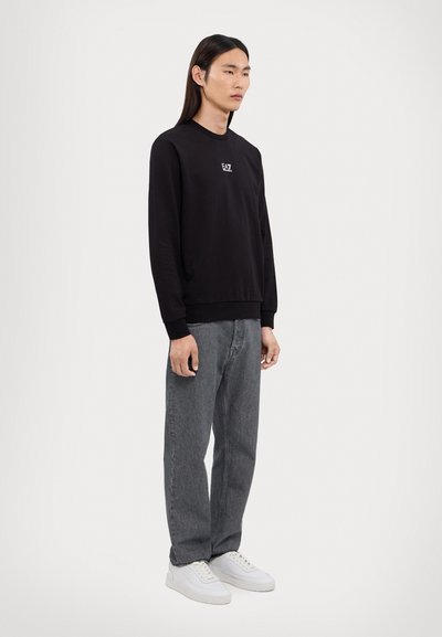 Sweatshirt noir avec des poignets et un ourlet côtelés, arborant un petit logo. Assorti avec un jean droit gris et des baskets blanches. Design minimaliste.