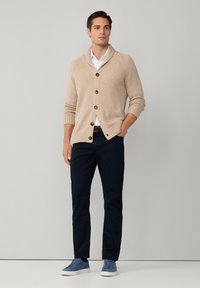 Cárdigan de punto beige con cuello chal y botones de madera, combinado con pantalones marinos y zapatillas azules. Textura suave, corte relajado.