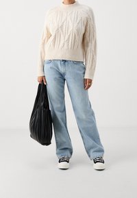 Personne portant un pull en maille torsadée crème, un jean ample bleu clair, des baskets noires et blanches, tenant un sac fourre-tout noir plissé sur le côté.