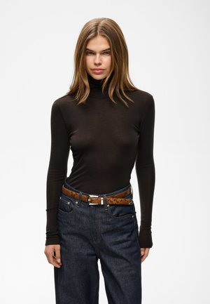 Jeune femme aux cheveux lisses châtain clair porte un col roulé noir à manches longues et un jean en denim foncé avec une ceinture marron, debout devant un fond blanc.