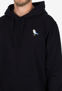 Sudadera azul marino de algodón con un bolsillo frontal. Presenta un pequeño diseño de pájaro bordado con acentos en gris y azul.