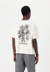 Camiseta blanca de algodón con un diseño floral en arte lineal negro en la parte trasera. Cuello redondo, mangas cortas, corte relajado, dobladillo estándar.