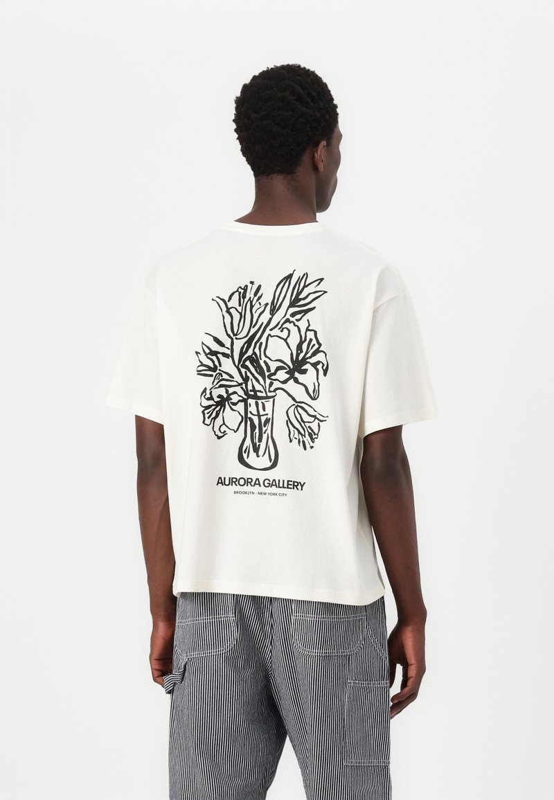 Camiseta blanca de algodón con un diseño floral en arte lineal negro en la parte trasera. Cuello redondo, mangas cortas, corte relajado, dobladillo estándar.