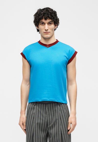 Jeune homme aux cheveux noirs bouclés portant un haut bleu sans manches avec des bordures rouges et un pantalon taille haute à rayures noires et blanches.