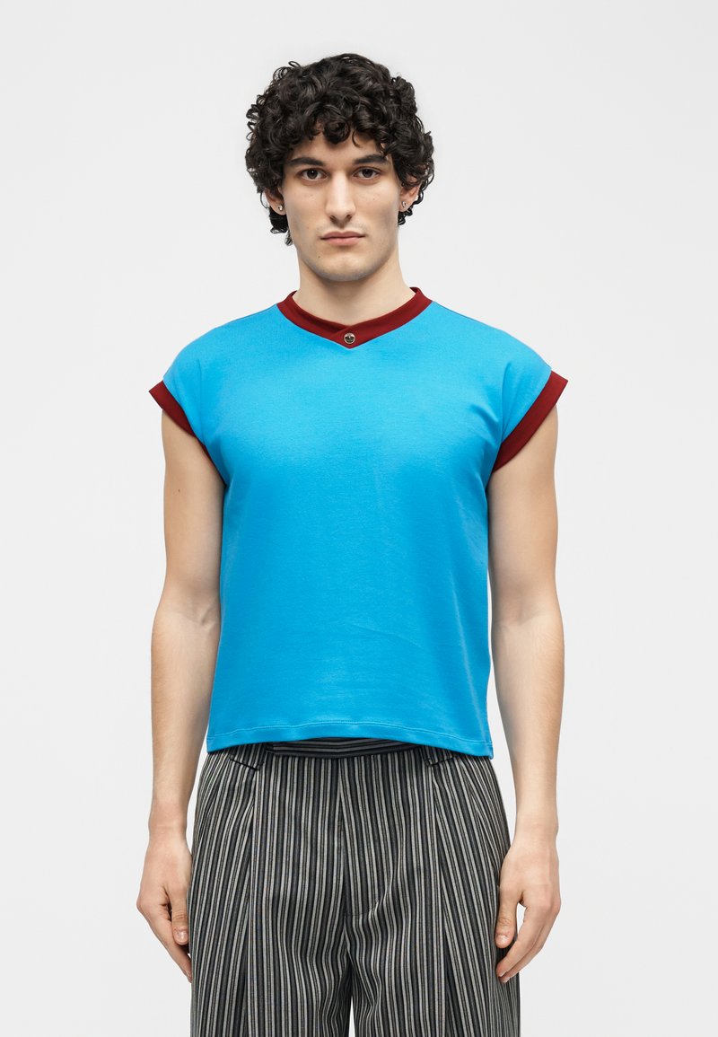 Young man with curly black hair wearing a sleeveless blue top with red trim and high-waisted black and white striped pants.