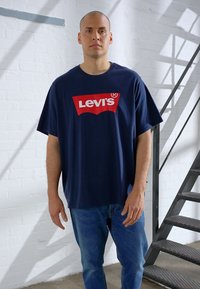 Sötétkék, nagyméretű póló piros Levi's logóval. Világoskék farmerrel párosítva. A modell egy textúrált fehér fal és lépcső mellett áll.