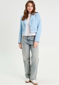 Veste courte texturée bleu clair avec un col et deux boutons argentés, portée sur une chemise blanche et associée à un jean en denim clair.