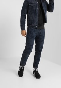 Jeansjacka och kamouflagebyxor i mörka nyanser, tillsammans med svarta träningsskor. Funktioner inkluderar knappstängning och slim fit-design.
