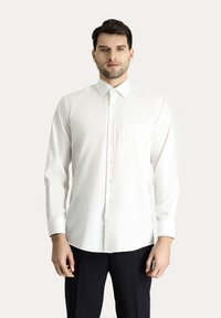 Camicia bianca con colletto classico, maniche lunghe e taschino sul petto. Tessuto liscio con vestibilità regolare. Indossata con pantaloni scuri.