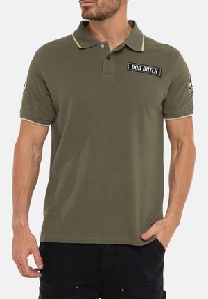 Homme portant un polo Von Dutch vert olive avec des écussons logo sur la poitrine et les manches, associé à un pantalon noir.