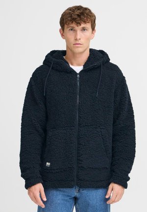 Sort fleece hoodie med lynlås foran, snor i hætten og to frontlommer. Tekstureret stof og afslappet pasform. Bæres over en hvid t-shirt.