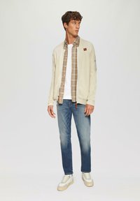 Helles beiges Zip-Up Cardigan über einem braun karierter Hemd, weißem T-Shirt und blauen Jeans; ergänzt mit cremefarbenen Turnschuhen.