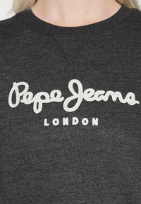 Sweat-shirt gris foncé avec texte blanc brodé "Pepe Jeans London" sur la poitrine, porté par une personne à la peau claire et aux cheveux blonds.
