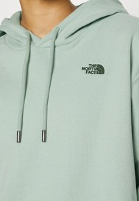 Felpa verde menta realizzata in morbido tessuto, con una tasca frontale, cordini con punte in metallo e un piccolo logo verde sul petto.