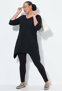 Zwarte tuniek met een V-hals en een asymmetrische zoom, gecombineerd met zwarte leggings. Het model draagt ronde brillen en houdt een witte tas vast. Getextureerde flat shoes.