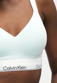 Žena nosi svetlomodrý bralette Calvin Klein s bielym elastickým pásom a čiernym logom, zobrazuje hornú časť trupu a plecia.