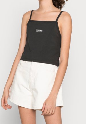 Kobieta ubrana w czarny top na ramiączkach Levi's i białe szorty z wysokim stanem, stojąca z luźno opuszczonymi ramionami na jednolitym tle.