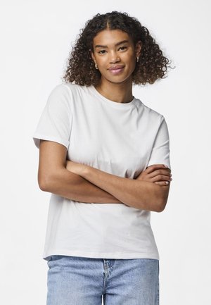 Hvit bomulls t-skjorte med korte ermer, rund hals og en avslappet passform, parret med lyseblå denim jeans. Modell med armene i kryss.