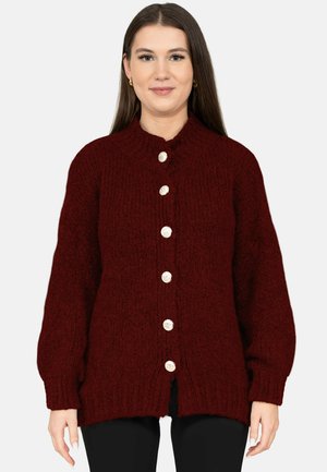 Vrouw draagt een donkerrode grofgebreide cardigan met zes grote zilveren knopen, gecombineerd met een zwarte broek, staand tegen een effen achtergrond.