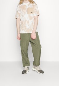 Kortärmad beige tie-dye t-shirt med bröstficka; gröna avslappnade byxor; grå skor med accentfärger och snörning. Nike-logga synlig.