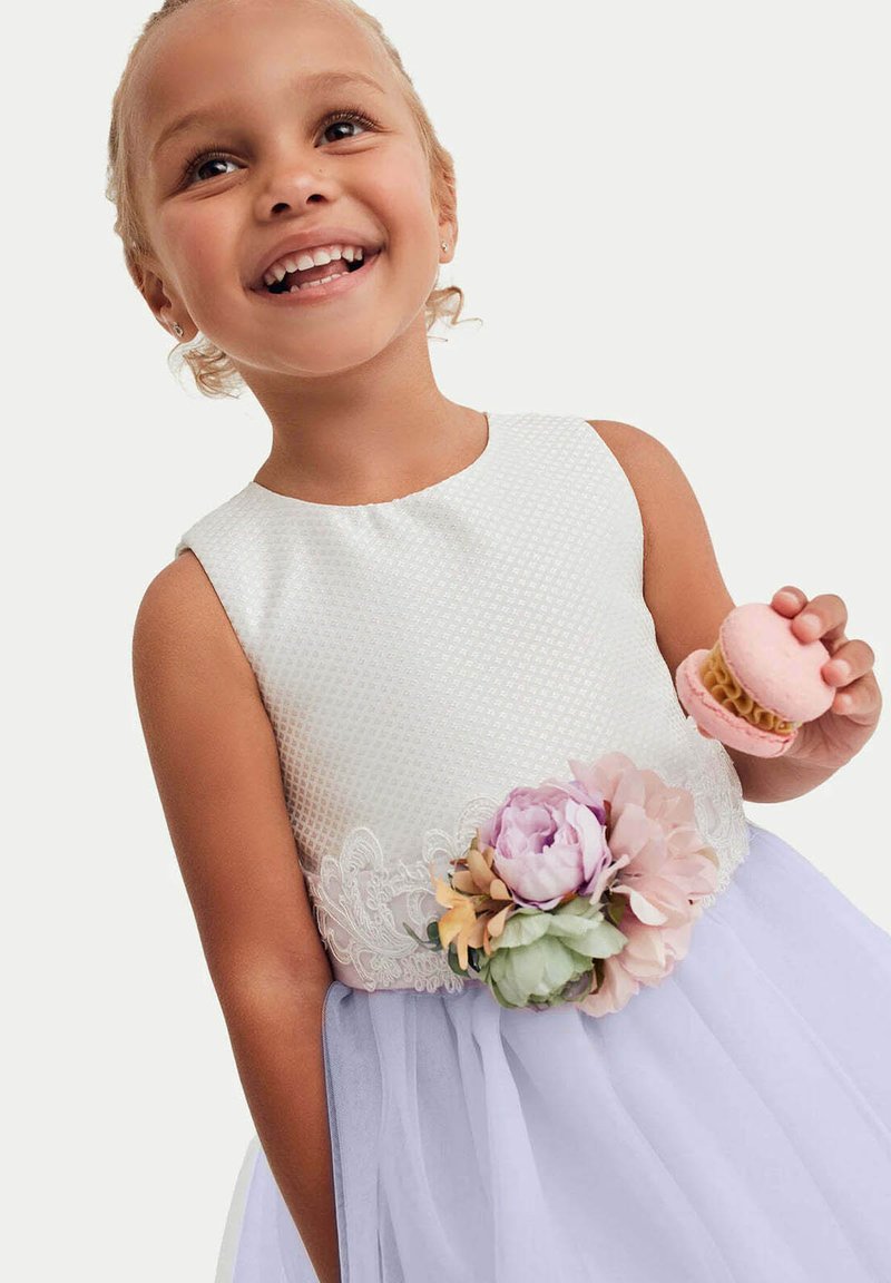 Abito bianco senza maniche con corpino texture e accentuazioni floreali; gonna in tulle lavanda; ragazza che tiene un macaron rosa.