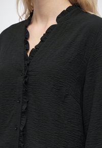 Gros plan d'un chemisier noir texturé avec de petits volants le long du col en V boutonné, porté par une personne à la peau claire et aux cheveux blonds.