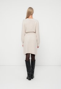 Stickad beige klänning med lång ärm och midjebälte, med rundad halsringning och en hög-låg fåll. Kombinerad med svarta knähöga stövlar.