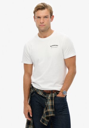 Hvid bomulds t-shirt med et lille sort "SUPERDRY BRITISH DESIGN" logo. Båret med mørke jeans og en blå ternet skjorte knyttet om taljen.